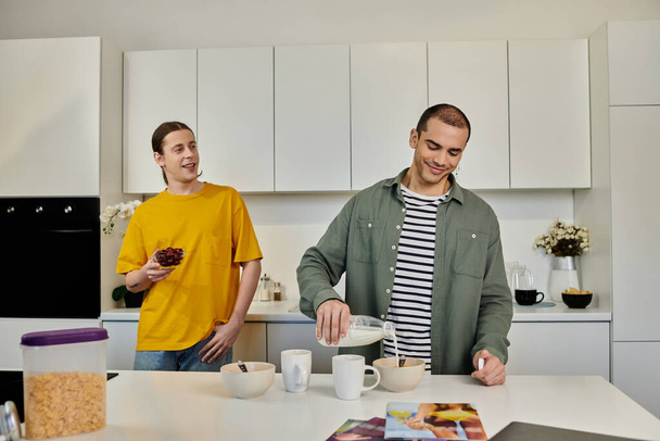 Ein junges schwules Paar frühstückt gemeinsam in seiner modernen Wohnung. Ein Mann gießt Milch in eine Schüssel, während der andere lächelnd zusieht. - Foto, Bild