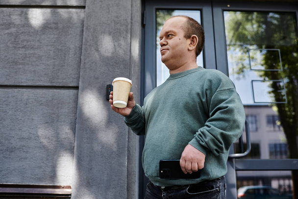 Um homem com inclusividade em uma camisola verde segura uma xícara de café enquanto olha para a esquerda. - Foto, Imagem