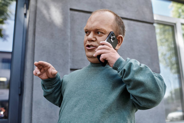 A man with inclusivity in a teal sweater talks animatedly on his phone outside a building. - Photo, Image