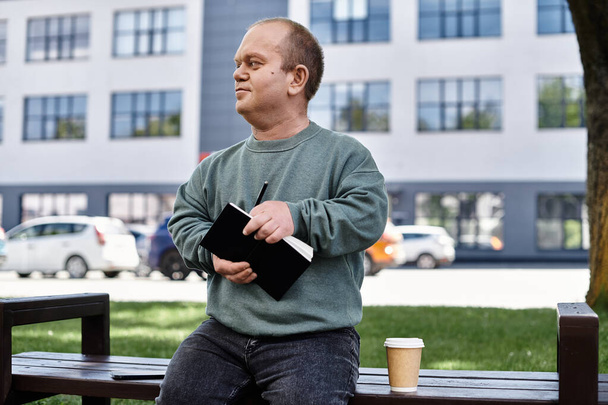 Een man met inclusiviteit zit op een bankje in een stadspark, bedachtzaam schrijvend in een notitieboekje met een koffie naast zich. - Foto, afbeelding