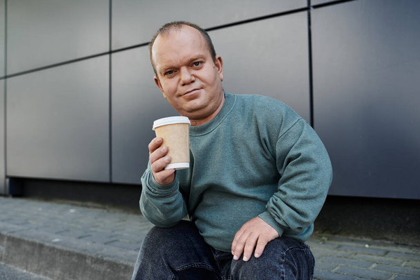 Un homme avec inclusivité est assis sur un trottoir, tenant une tasse de café et souriant pour la caméra. - Photo, image
