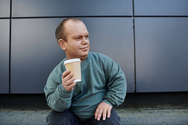 Muž s inkluzivitou a šálkem kávy se na chvíli odmlčí a dívá se na město.. - Fotografie, Obrázek