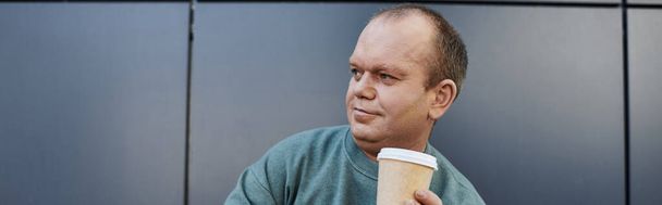 Ένας άντρας με εγκλειστικότητα και στοχαστική έκφραση κρατάει ένα φλιτζάνι καφέ, απολαμβάνοντας μια στιγμή ηρεμίας στην πολυάσχολη μέρα του.. - Φωτογραφία, εικόνα