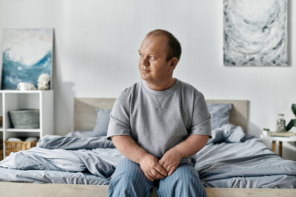Een man met inclusiviteit zit aan de rand van een bed in een slaapkamer, op zoek bedachtzaam. - Foto, afbeelding