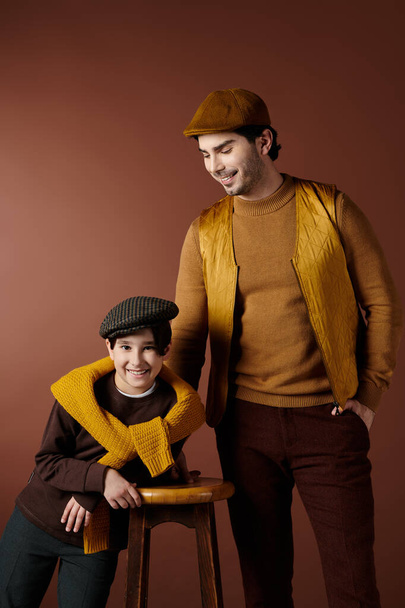 A father and son stand together in a studio setting, both wearing warm brown and yellow clothing. They are smiling and appear to be enjoying each others company. - 写真・画像