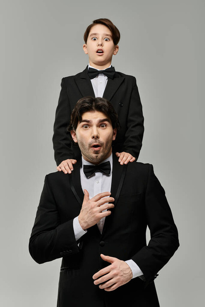 Um pai e um filho em smoking combinando, ambos parecendo surpresos, com o filho em pé sobre os ombros dos pais. - Foto, Imagem