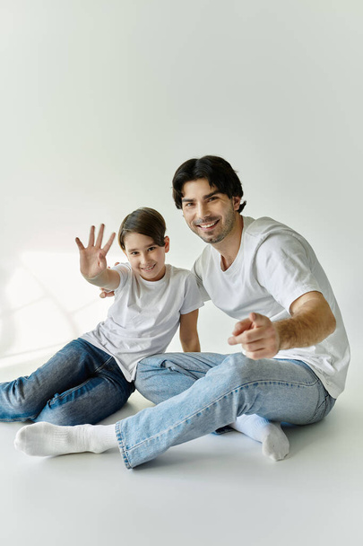 Isä ja poika istuvat yhdessä valkoisella lattialla hymyillen ja vilkuttaen kameralle osoittaen läheistä sidettään.. - Valokuva, kuva