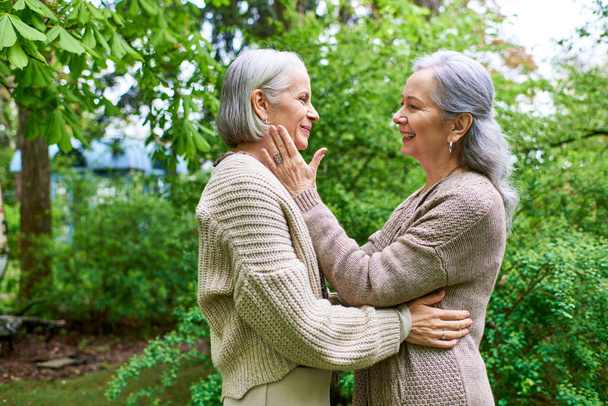 Δύο μεσήλικες γυναίκες, ντυμένες με ζακέτες, αγκαλιάζονται με αγάπη σε ένα καταπράσινο περιβάλλον κήπου.. - Φωτογραφία, εικόνα