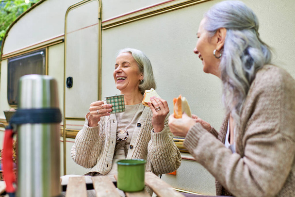 Két középkorú nő élvezi a kávészünetet, miközben az erdőben kempingeznek, nevetnek és együtt esznek.. - Fotó, kép