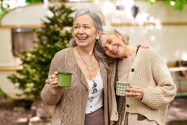 Ein lesbisches Paar genießt eine Tasse Kaffee zusammen, während es in einem sattgrünen Wald zeltet. Sie lächeln und lachen, genießen ihre gemeinsame Zeit. - Foto, Bild