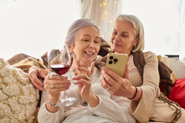 Dwie kobiety w średnim wieku relaksują się w kampingu, delektując się lampką wina i sprawdzając smartfona - Zdjęcie, obraz