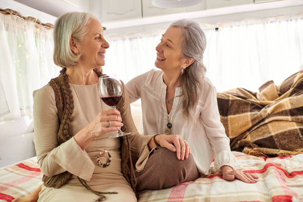 Dos mujeres de mediana edad disfrutan de una copa de vino en su acogedora caravana durante un viaje por carretera. - Foto, imagen