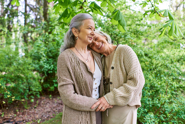 Due donne di mezza età che indossano cardigan si abbracciano in una lussureggiante foresta verde, godendo di un momento di vicinanza e affetto. - Foto, immagini