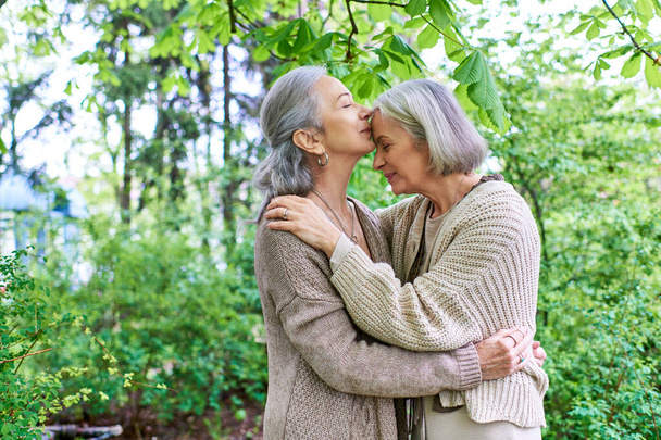 Ein lesbisches Paar in Strickjacken umarmt sich zärtlich im Schatten eines grünen Baumes, ihre Liebe ist in ihrer Umarmung spürbar. - Foto, Bild