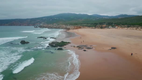 Beautiful ocean foaming on sandy coast with coastal cliffs in super slow motion. Frothy waves washing picturesque beach cloudy day aerial view. Green mountains tower on gray horizon at background. - Footage, Video