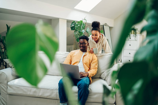 Feliz joven pareja multirracial viendo vídeo en un ordenador portátil juntos. Hombre afroamericano sonriente sentado en un sofá y mujer hispana de pie detrás de él con una taza de café o té en la mano. - Foto, imagen