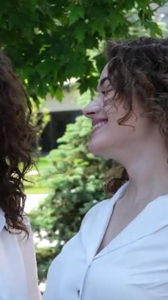 Στο πάρκο σε λευκές μπλούζες Δύο γυναίκες μητέρα και κόρη γελούν χαμογελώντας παρόμοια σγουρά κορίτσια. Κοιτάξτε ο ένας τον άλλον - Πλάνα, βίντεο