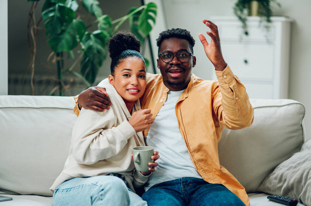 ハッピーヒスパニック系の女性とアフリカ系アメリカ人男性が,コーヒーを飲んで抱擁しながらテレビを見ています. 一緒に映画や映画の夜を楽しんでいる笑顔の多民族カップル. - 写真・画像