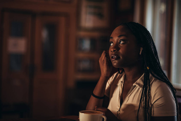 Bella giovane donna afro-americana che si mette gli airpoods nelle orecchie mentre è seduta in un caffè e beve caffè - Foto, immagini