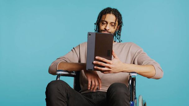 Homem em cadeira de rodas conversando com amigo através de videocall internet usando tablet. Pessoa com deficiência segurando dispositivo portátil, discutindo com o companheiro em teleconferência, fundo do estúdio, câmera B - Foto, Imagem