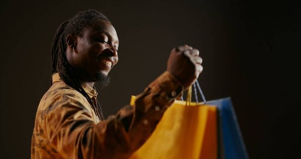 Happy pleased person carrying shopping bags after buying favorite clothes on discount, enjoying sales season at the mall. African american shopaholic guy looks at his new clothing items in bags. - 写真・画像