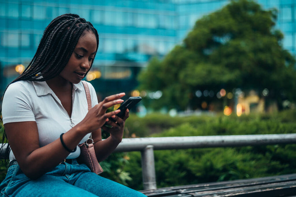 Vrolijk Afrikaans-Amerikaanse vrouw met behulp van smartphone terwijl zitten op de bank in de stad - Foto, afbeelding