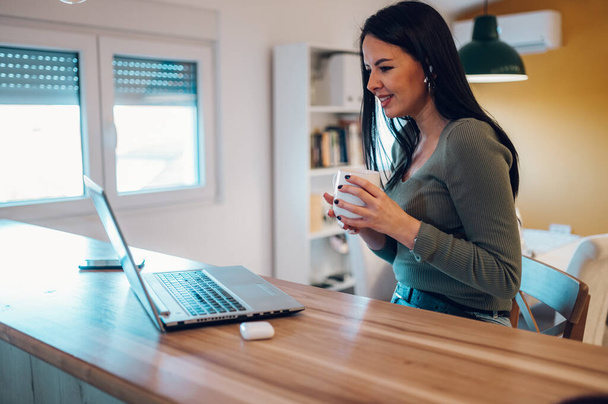 Piękna kobieta w średnim wieku pijąca poranną kawę i patrząca na monitor laptopa w kuchni w domu. Cieszy się poranną kawą. - Zdjęcie, obraz