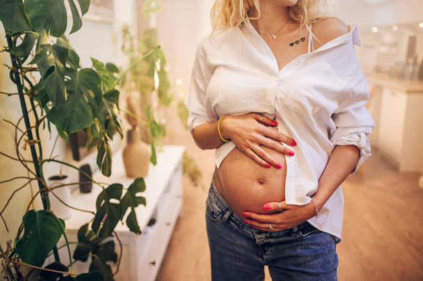 Schnappschuss einer schwangeren Frau, die in ihrem Wohnzimmer steht und ihren hübschen Bauch zeigt. Attraktive werdende Mutter lächelt und posiert zu Hause und hält die Hände auf dem Bauch. - Foto, Bild