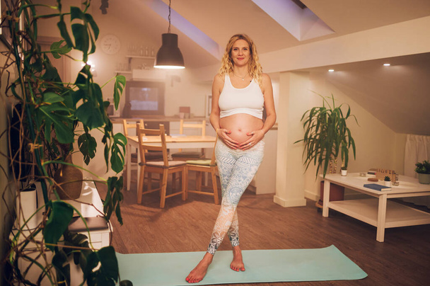 Schöne schwangere Frau, die auf der Matte steht und ihren Bauch berührt, nachdem sie pränatale und meditative Yoga-Übungen durchgeführt hat. Pränatale Übungen. Kopierraum. - Foto, Bild