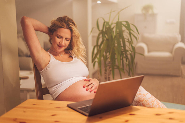 Hamile kadın evde dizüstü bilgisayar kullanıyor. Müstakbel anne mutfak masasında otururken bilgisayar kullanıyor. Arkadaşlarla ve ailelerle iletişim, online alışveriş ya da hamilelik sırasında iş. - Fotoğraf, Görsel