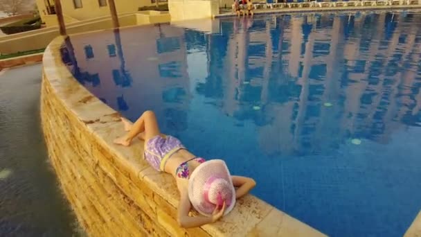 top view of a young girl in swimwear lying down by a serene resort swimming infinity pool, enjoying a sunny day with a pink hat beside her on Dead Sea view - Footage, Video
