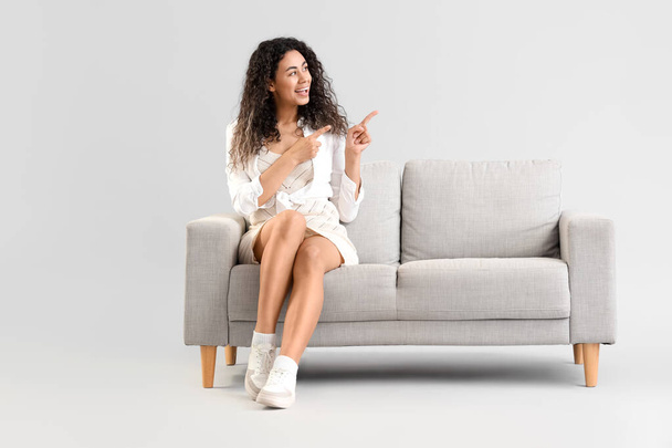 Giovane donna afro-americana sul divano grigio che punta a qualcosa sullo sfondo chiaro - Foto, immagini