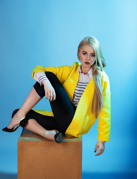 Portrait of blond model in fashion yellow coat sitting over blue - Foto, afbeelding