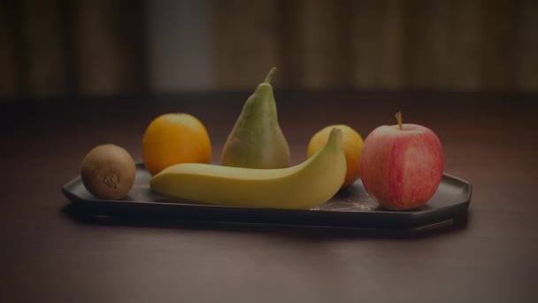 Een selectie van vruchten, waaronder een banaan, peer, sinaasappel, appel en kiwi worden weergegeven op een dienblad op een tafel, klaar om te genieten. Deze natuurlijke voedingsmiddelen zijn nietjes in een gezond dieet - Video