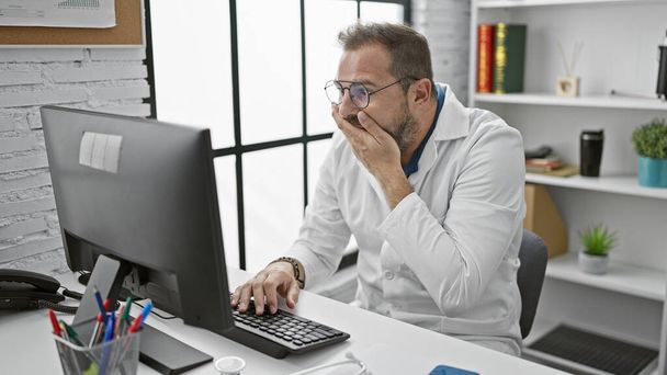 Egy meglepett spanyol férfi ősz hajjal fehér köpenyben számítógépen dolgozik egy klinika irodájában.. - Fotó, kép
