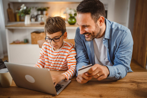 Padre adulto caucásico hombre mantener la tarjeta de crédito, mientras que el hijo chico comprar en línea en el ordenador portátil en la cocina - Foto, Imagen