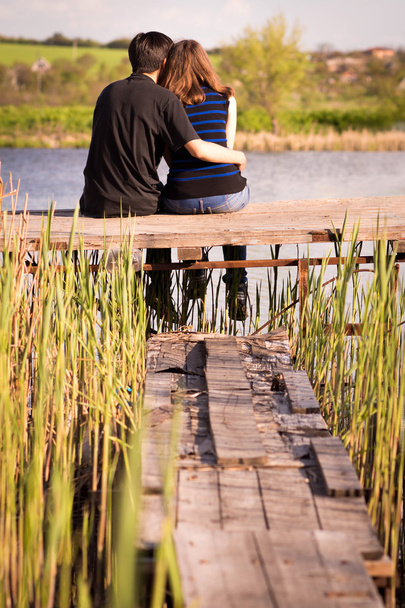Мужчина обнимает девушку, сидящую на пирсе на берегу реки
 - Фото, изображение