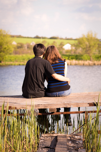 άνδρας αγκαλιάζει το κορίτσι που κάθεται σε μια προβλήτα στην όχθη ποταμού - Φωτογραφία, εικόνα