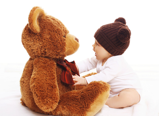 Bebê de malha chapéu marrom brincando com grande ursinho de pelúcia
 - Foto, Imagem