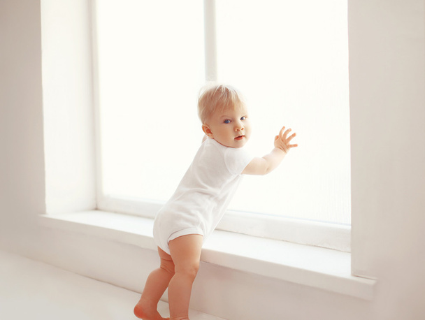 Little baby at home in white room stands near window - Φωτογραφία, εικόνα