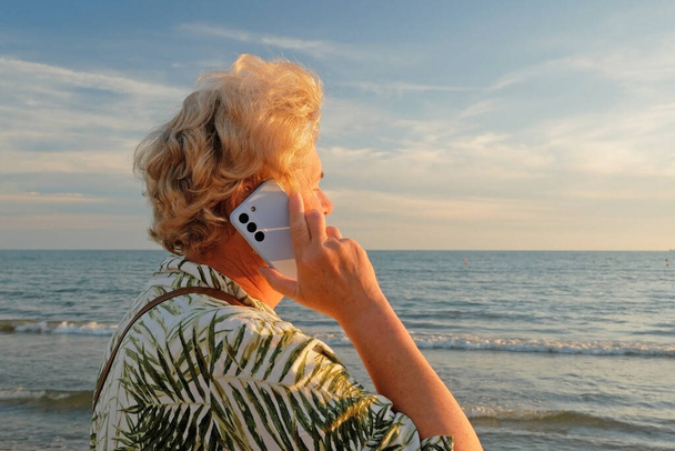 Femme âgée séjournant sur la plage de la mer et parlant avec un téléphone portable dans les rayons du coucher du soleil. Femme mûre sourit tenant le téléphone portable dans la main. Concept d'itinérance, wifi, vacances, liberté et retraite heureuse. À l'extérieur - Photo, image