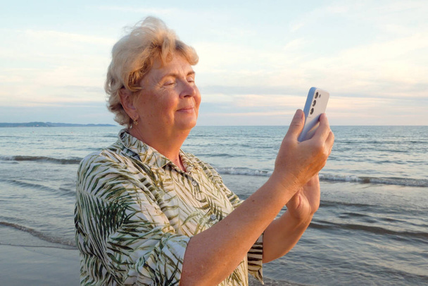 Femme âgée séjournant sur la plage de la mer et parlant avec un téléphone portable dans les rayons du coucher du soleil. Femme mûre sourit tenant le téléphone portable dans la main. Concept d'itinérance, wifi, vacances, liberté et retraite heureuse. À l'extérieur - Photo, image