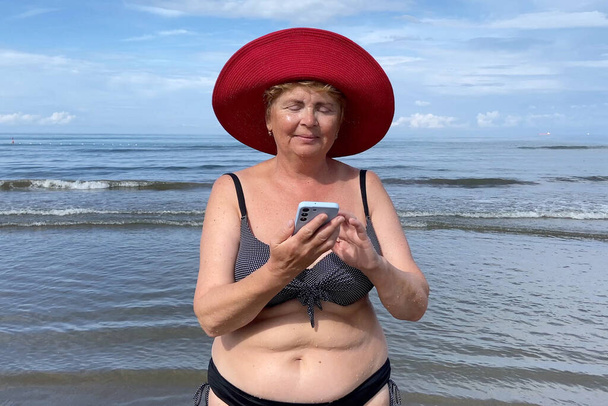 Femme âgée portant un maillot de bain séjournant sur la plage de la mer et parlant avec un téléphone portable par temps ensoleillé. Femme mûre sourit tenant le téléphone portable dans la main. Concept d'itinérance, wifi, vacances, liberté et retraite heureuse. À l'extérieur - Photo, image