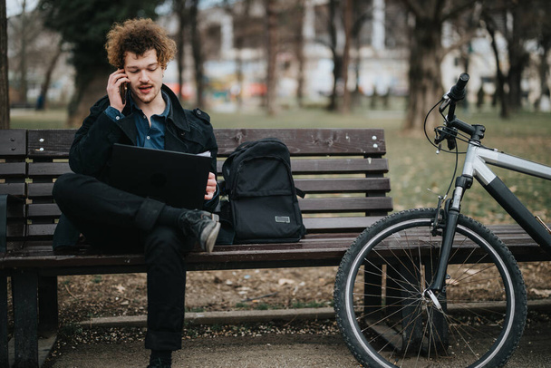 Nuori yrittäjä työskentelee kannettavan tietokoneen parissa ja puhuu puhelimessa istuessaan puistossa polkupyörän vieressä. - Valokuva, kuva
