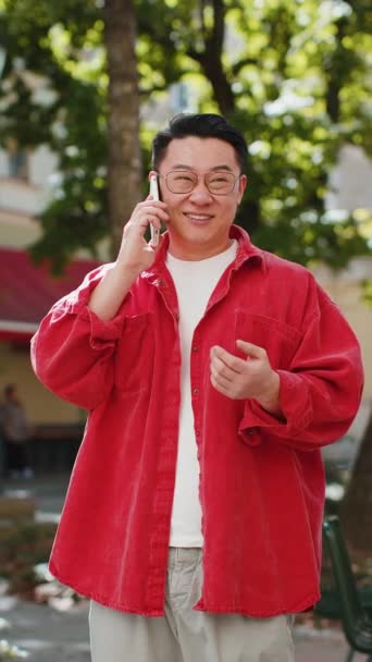 Szczęśliwy azjatycki mężczyzna w średnim wieku rozmawiający zdalnie przez smartfona na świeżym powietrzu. Japończyk rozmawiający przez telefon niespodziewane dobre wieści plotkują na ulicach miasta. Miejski styl życia. Pionowe - Materiał filmowy, wideo