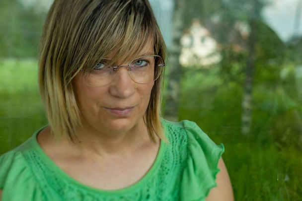 Μια γυναίκα με γυαλιά και μια πράσινη κορυφή ατενίζει με περίσκεψη μέσα από ένα παράθυρο, αντανακλώντας το πράσινο έξω. Η γαλήνια έκφραση και το φυσικό υπόβαθρο προσθέτουν μια ηρεμιστική επίδραση. - Φωτογραφία, εικόνα