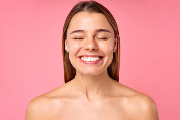 Het portret vangt een jonge vrouw op een levendige roze achtergrond, met gesloten ogen en een stralende glimlach die vreugde en positiviteit uitstraalt, geluk uitstraalt en juicht in een moment van gelukzaligheid - Foto, afbeelding