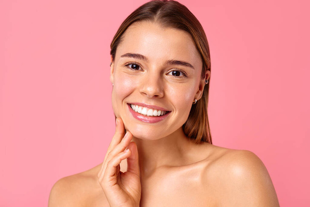 Eine fröhliche junge Frau strahlt vor rosa Hintergrund ein strahlendes Lächeln aus, das ihre klare, strahlende Haut und natürliche Schönheit zur Geltung bringt. Perfekt für Wellness-, Pflege- und Positivitätskonzepte. - Foto, Bild