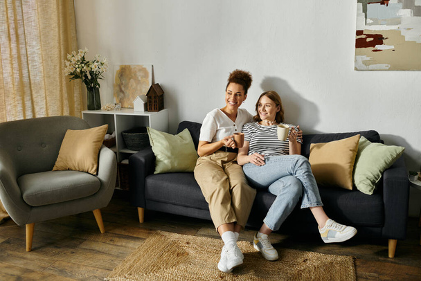 Zróżnicowana lesbijska para cieszy się relaksującym popołudniem w domu, dzieląc się kawą i uśmiechem na kanapie. - Zdjęcie, obraz