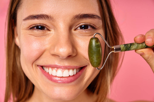 A woman on a pink background is smiling as she uses a jade roller for a facial massage, promoting a healthy, glowing complexion. This showcases selfcare and contemporary beauty practices - Photo, Image